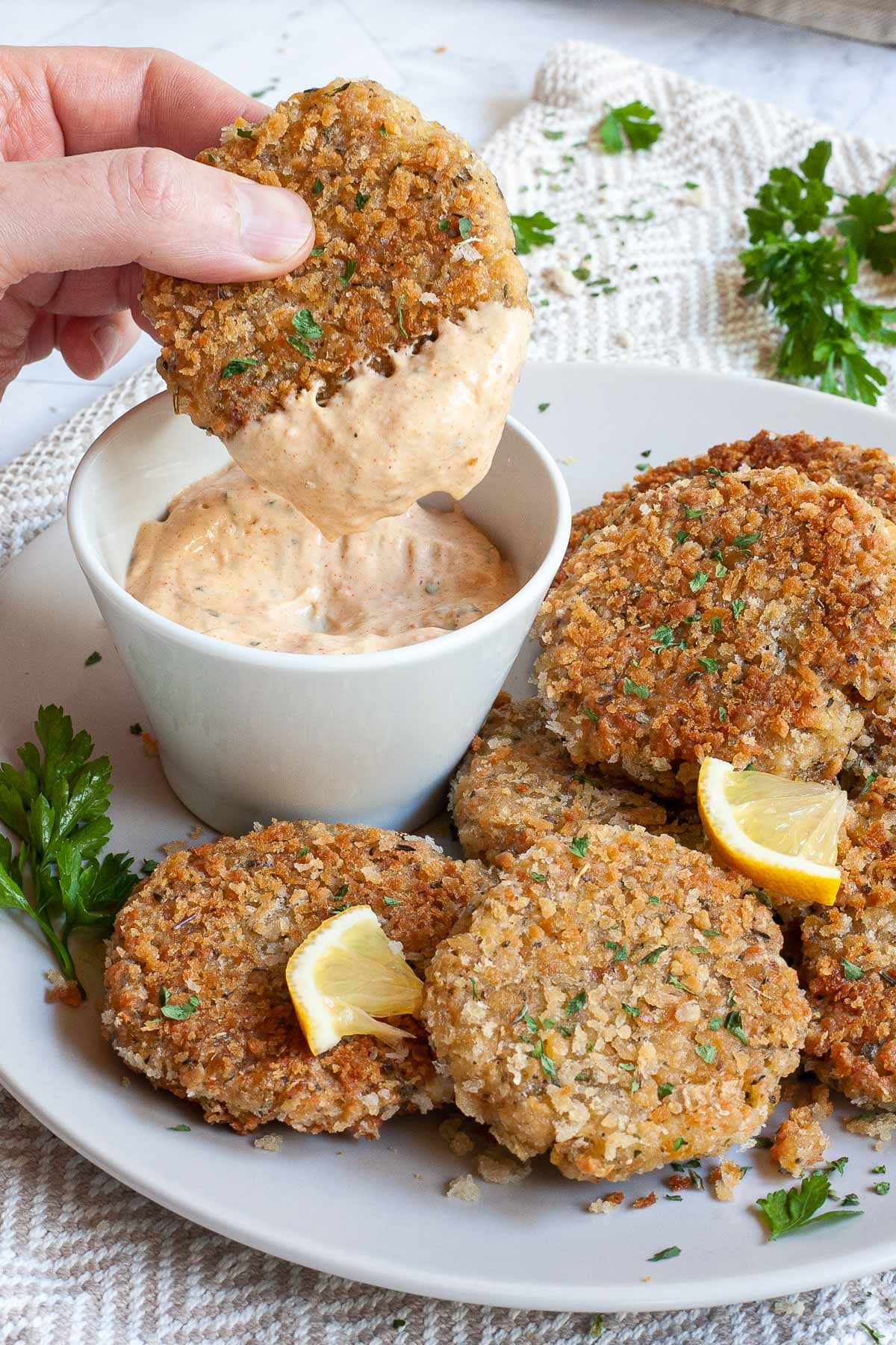 Several round-shaped breadcrumbs covered patties on a large white plate with a light orange sauce in a white bowl. Chopped parsley and lemon wedges are around for decoration. A hand is dipping one in the sauce.