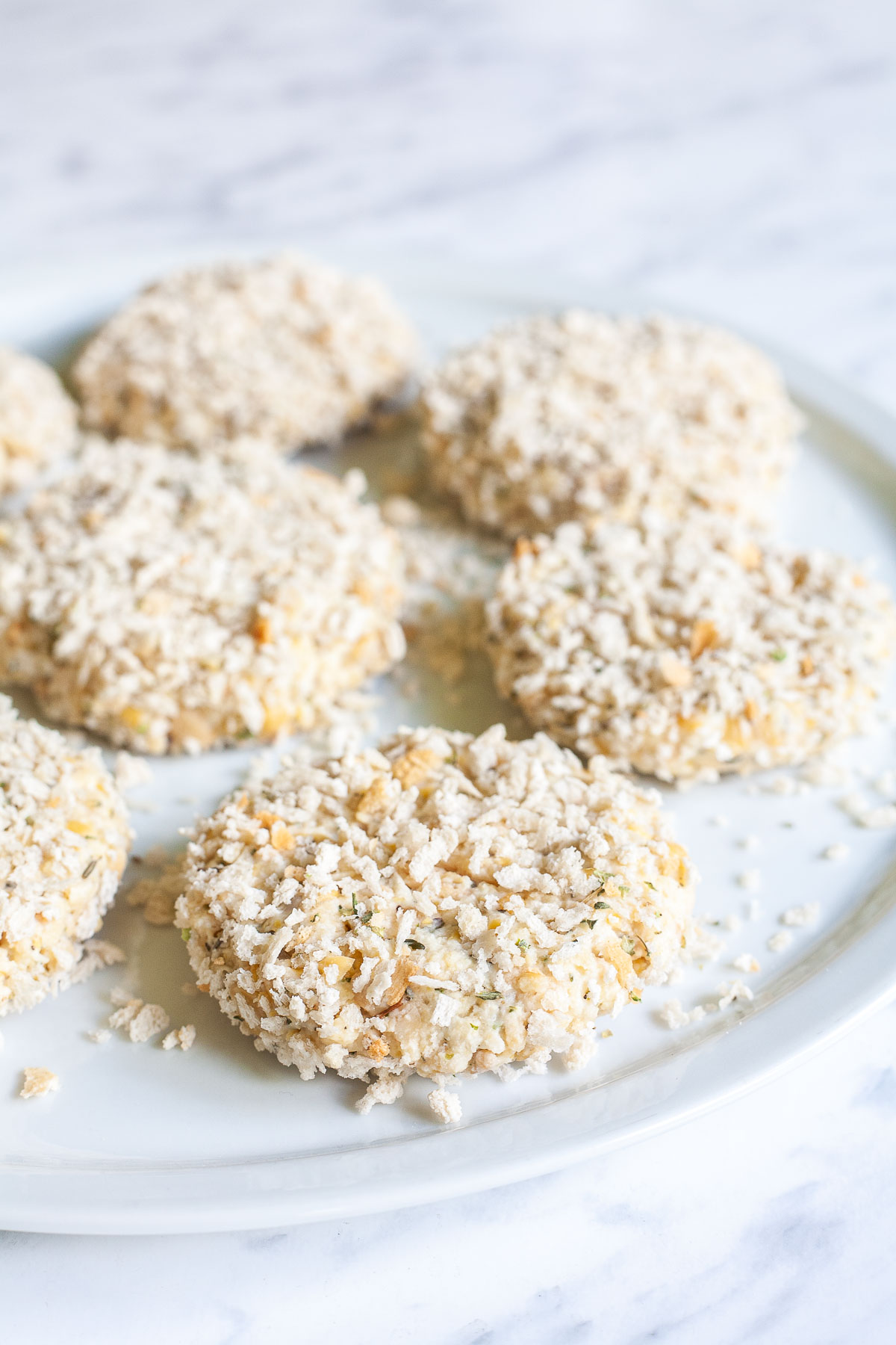 Several flat patties covered in panko breadcrumbs and placed on a white plate. 