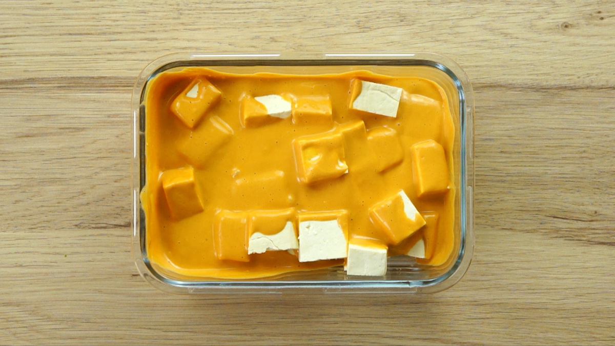 White raw tofu cubes in a thick orange marinade in a glass container.