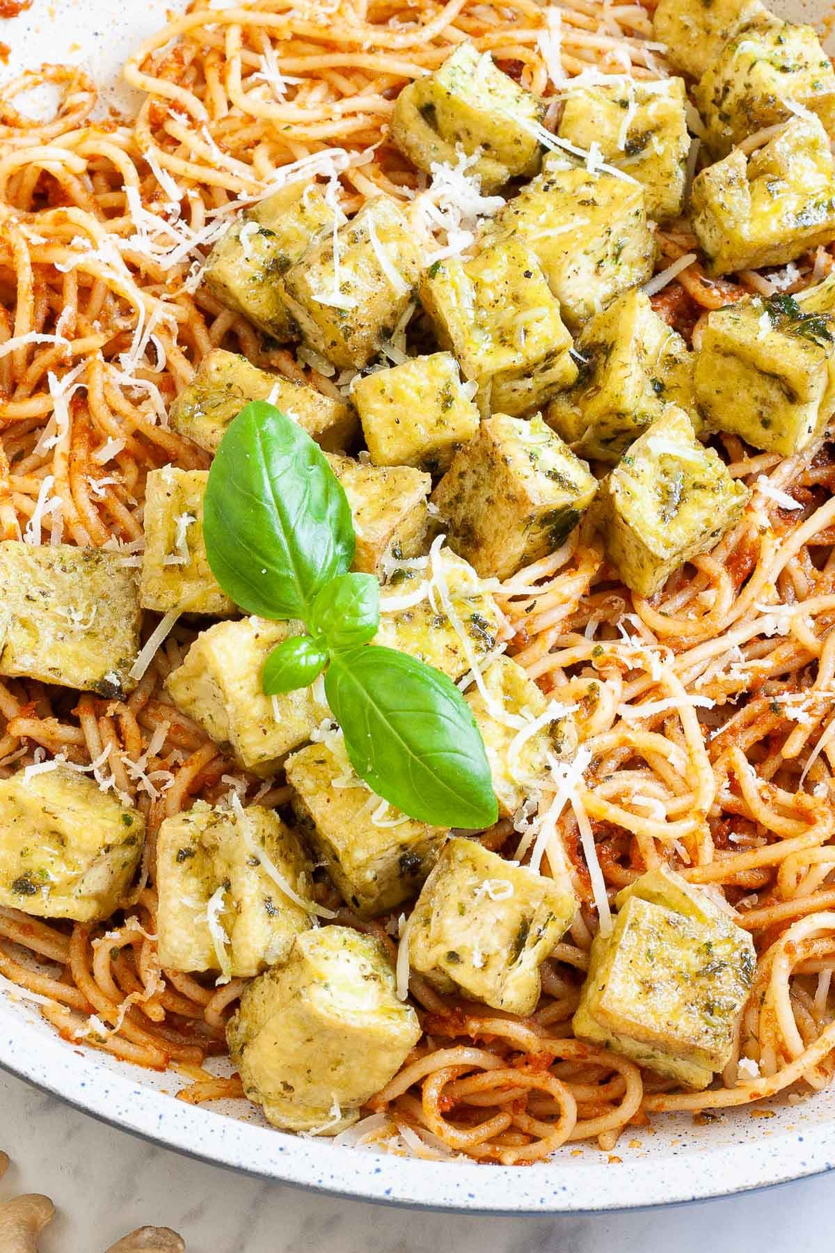 Large white frying pan with red pesto spaghetti topped with green-yellow tofu cubes, shredded cheese and basil leaves.