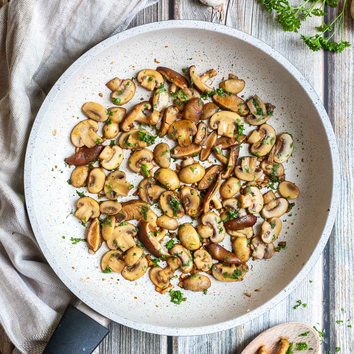 Golden brown sliced button and shiitake mushrooms in a frying pan topped with freshly chopped greens. A wooden spoon is next to it as well as some raw ingredients.