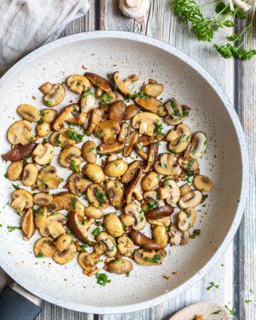 Golden brown sliced button and shiitake mushrooms in a frying pan topped with freshly chopped greens. A wooden spoon is next to it as well as some raw ingredients.