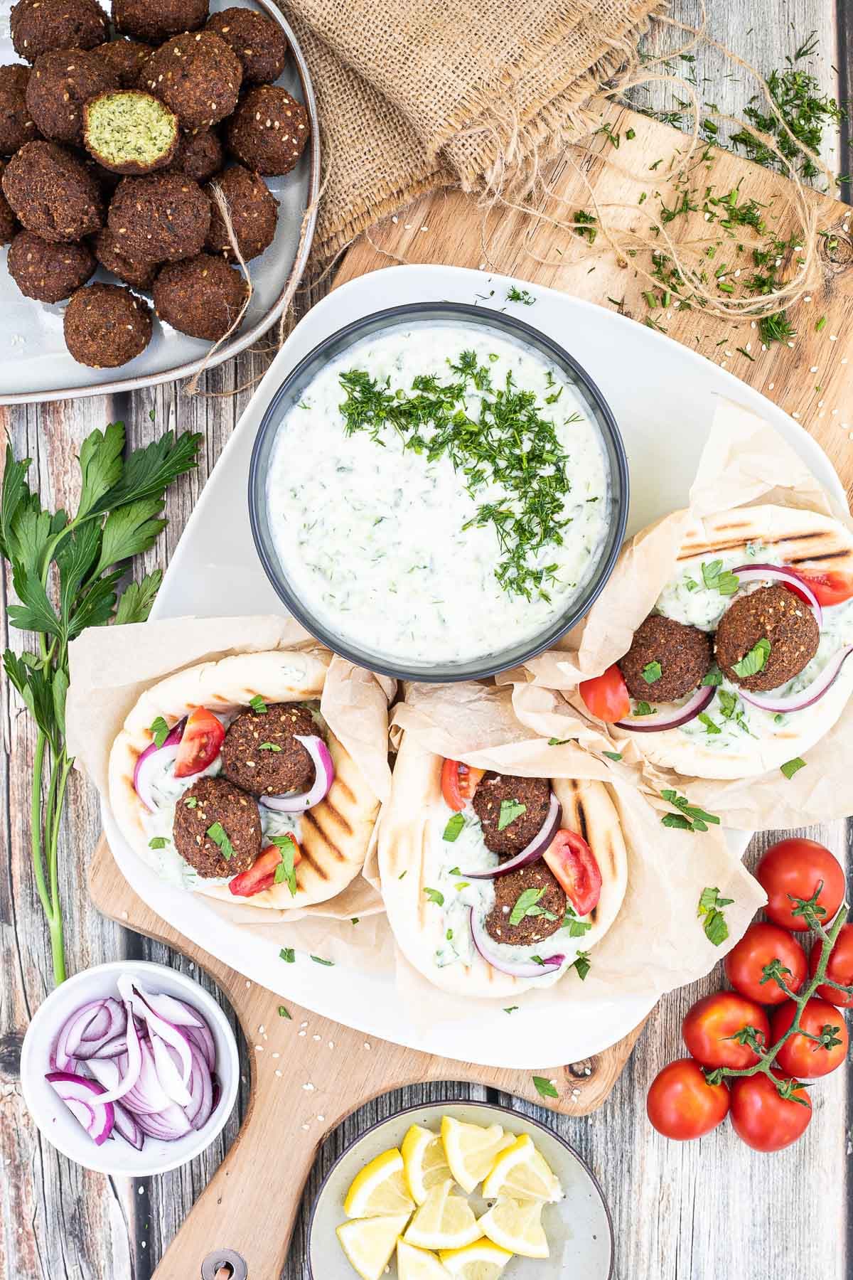 3 pitas folded and stuffed with 2 brown falafel balls, white grated cucumber salad, tomato and purple onion slices. A black bowl of white grated cucumber sauce is placed next to them. Leftover ingredients are placed in balls and on plates around it.