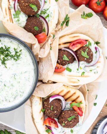 3 pitas folded and stuffed with 2 brown falafel balls, white grated cucumber salad, tomato and purple onion slices. A black bowl of white grated cucumber sauce is placed next to them.