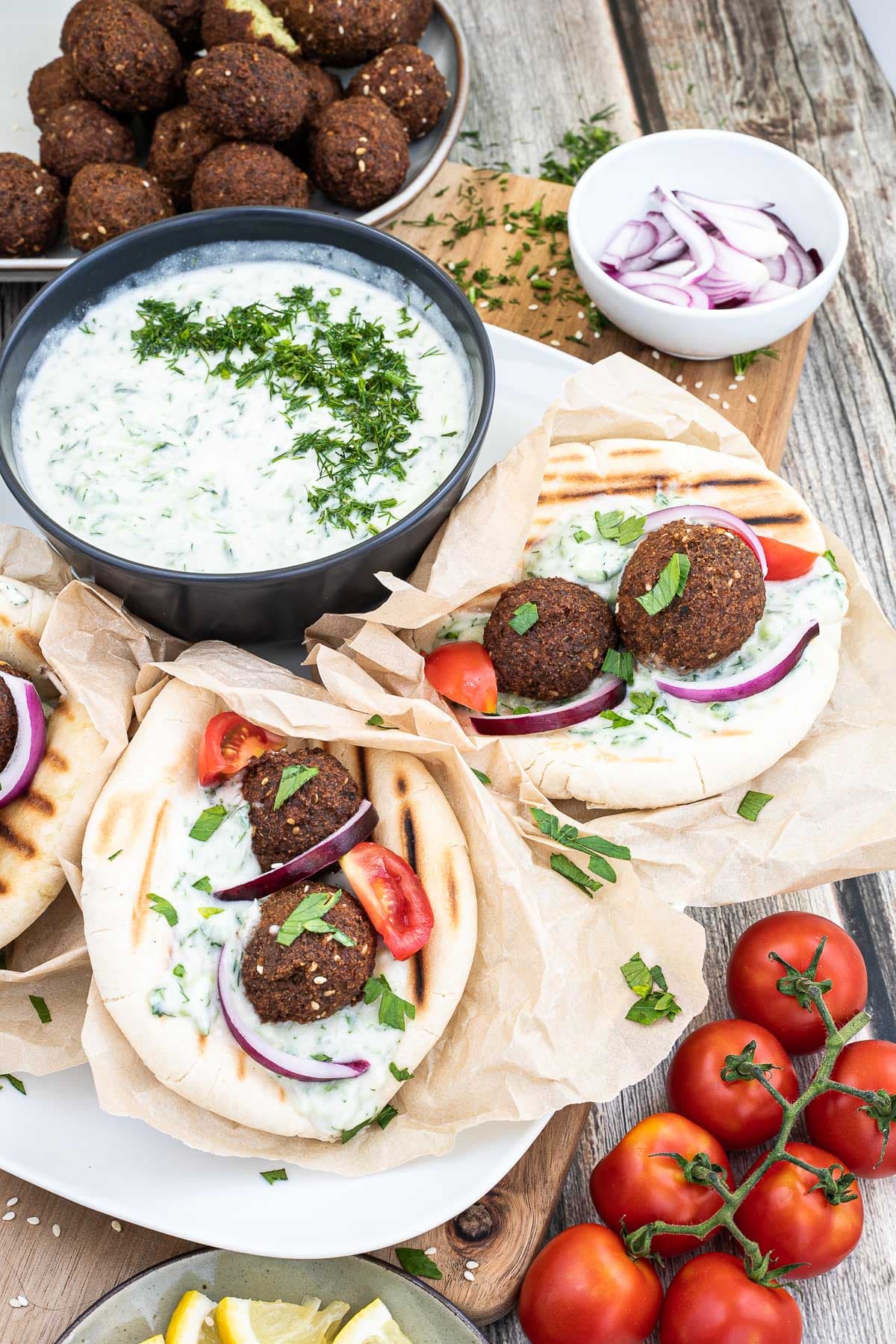 3 pitas folded and stuffed with 2 brown falafel balls, white grated cucumber salad, tomato and purple onion slices. A black bowl of white grated cucumber sauce is placed next to them. Leftover ingredients are placed in balls and on plates around it.