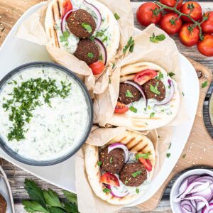 3 pitas folded and stuffed with 2 brown falafel balls, white grated cucumber salad, tomato and purple onion slices. A black bowl of white grated cucumber sauce is placed next to them.
