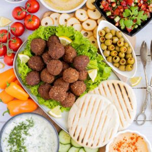 Lot of items in a white board including falafel balls on top of lettuce leaves, charcoaled pita breads, grated cucumber in yoghurt sauce, orange hummus, olives, chopped green red salad, light brown sauce, cherry tomatoes, orange peppers, sliced cucumber, breadsticks and crackers.