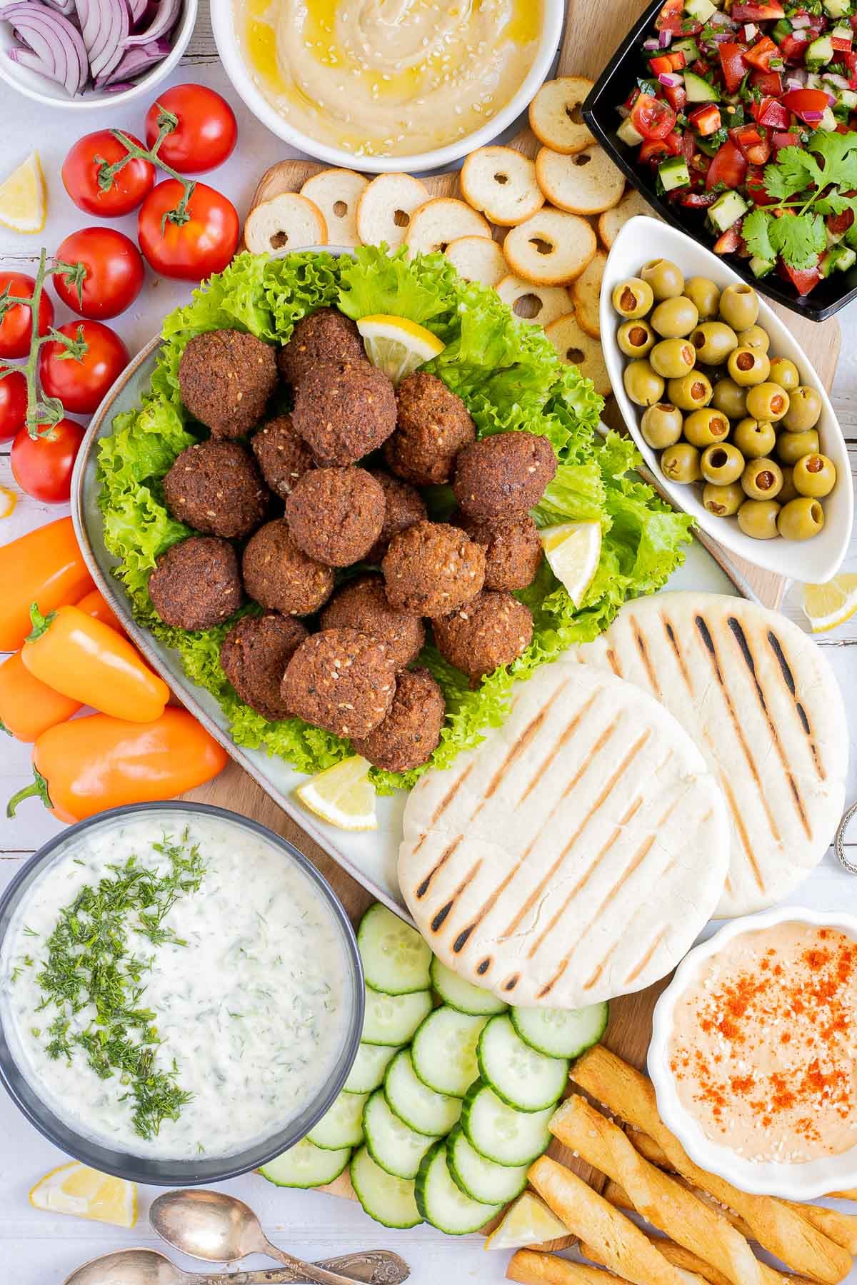 Lot of items in a white board including falafel balls on top of lettuce leaves, charcoaled pita breads, grated cucumber in yoghurt sauce, orange hummus, olives, chopped green red salad, light brown sauce, cherry tomatoes, orange peppers, sliced cucumber, breadsticks and crackers.