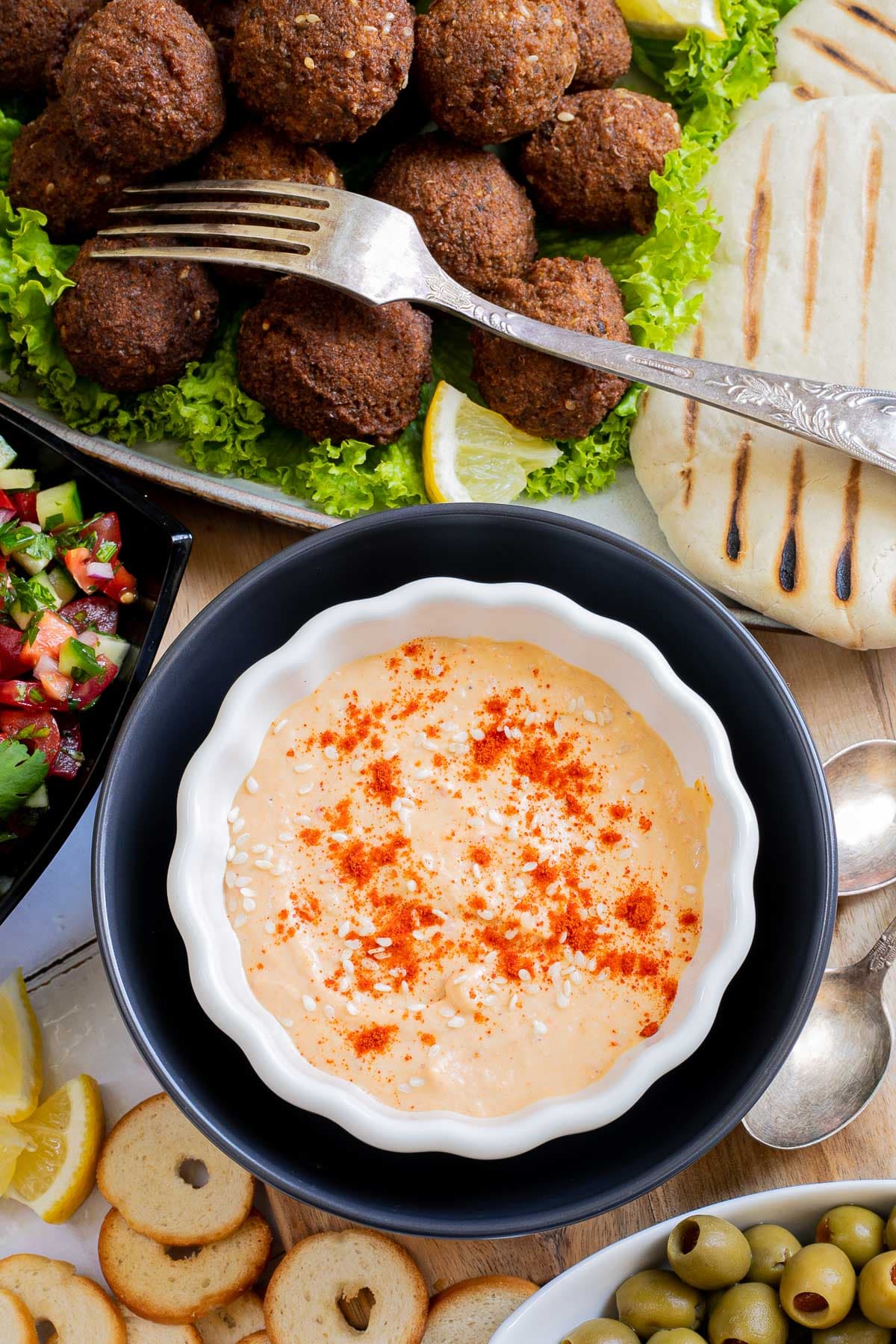 Orange hummus in white-black bowl. Falafel balls, pita breads, green olives, and chopped red green veggie salad is on the side.