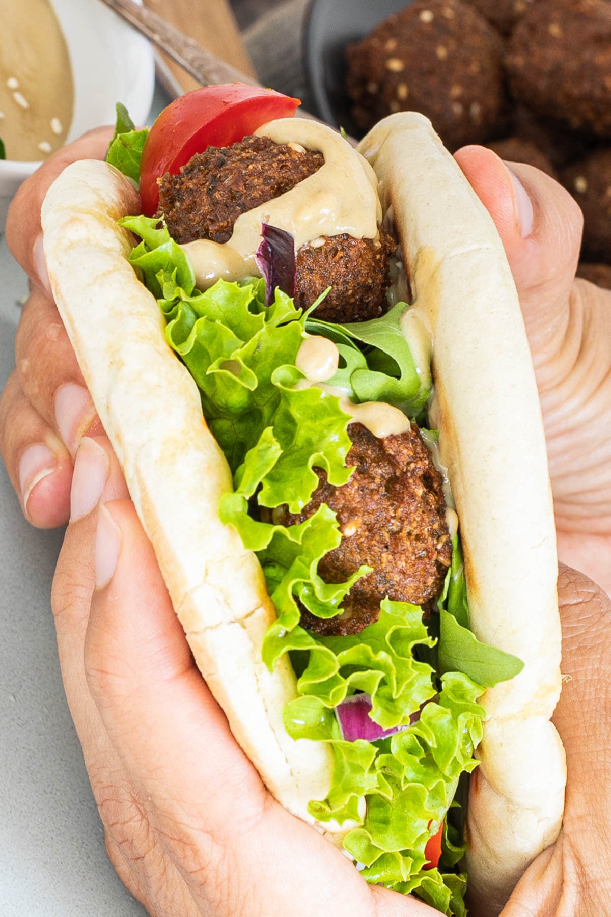 A hand is holding two pita breads as a sandwich filled with dark brown falafel bowls, vibrant green ruffled lettuce and light brown thick sauce.