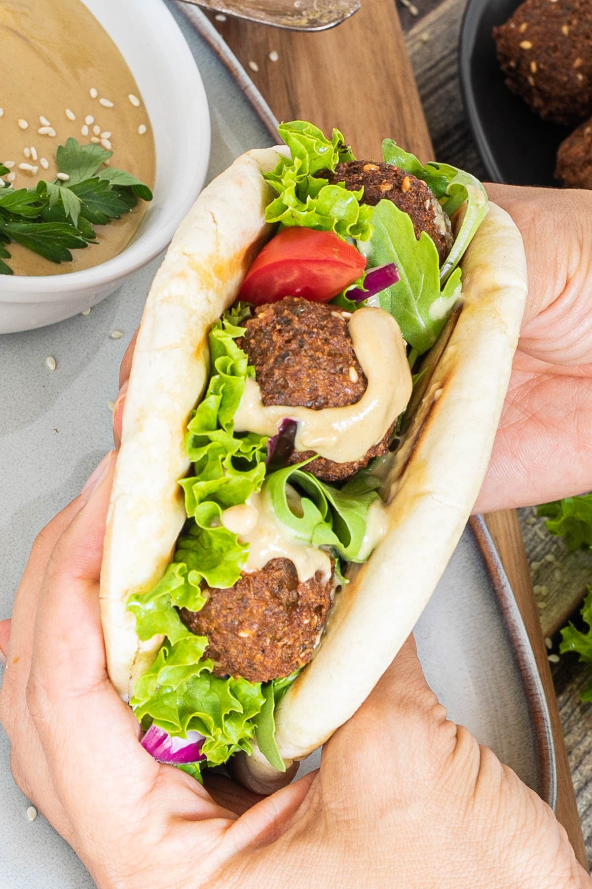 A hand is holding two pita breads as a sandwich filled with dark brown falafel bowls, vibrant green ruffled lettuce and light brown thick sauce.