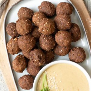 Falafel balls are served on a large plate. A light brown sauce in a white shallow bowl sprinkled with white sesame seeds and topped with a twig of parsley is next to them..
