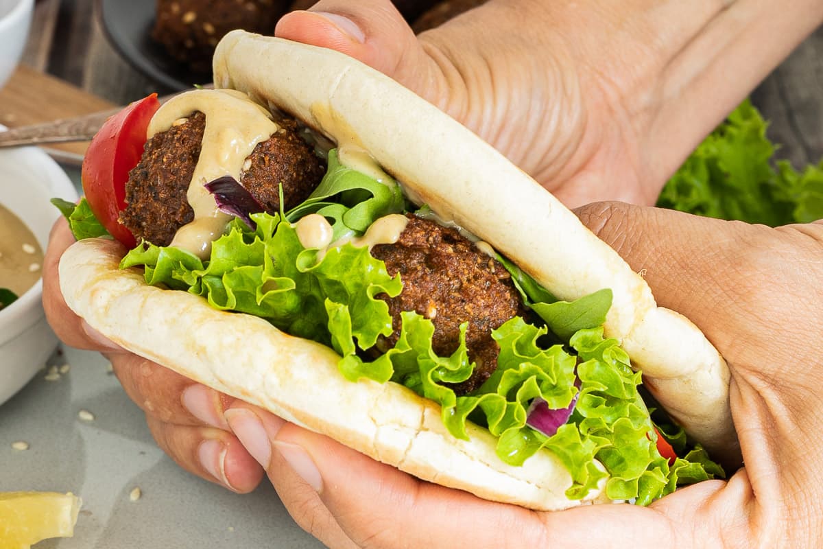 A hand is holding two pita breads as a sandwich filled with dark brown falafel bowls, vibrant green ruffled lettuce and light brown thick sauce.