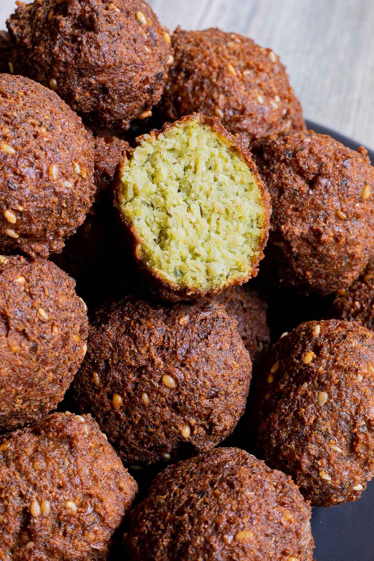 Dark brown falafel bowls on a black plate. One is cut in half and placed on to to see the green inside.