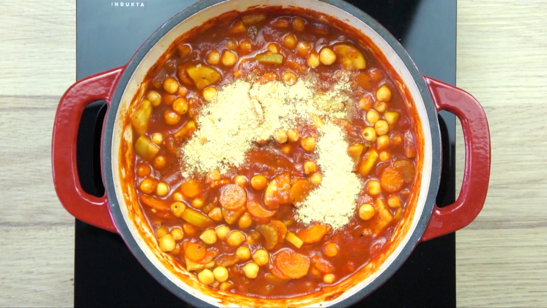 Veggie chunks and chickpeas covered with red tomato sauce and a small heap of yellow flakes in a white red enamelled Dutch oven.