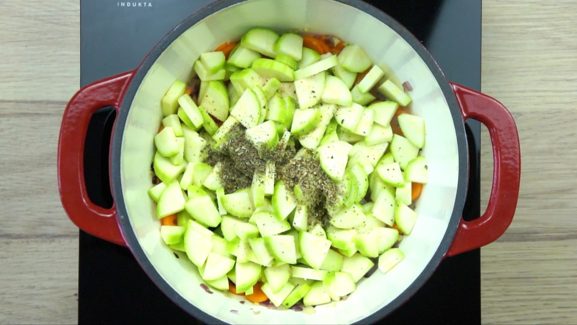 Chopped onion, garlic, zucchini, and sliced carrots with a small heap of dried green herbs in a white red enamelled Dutch oven.