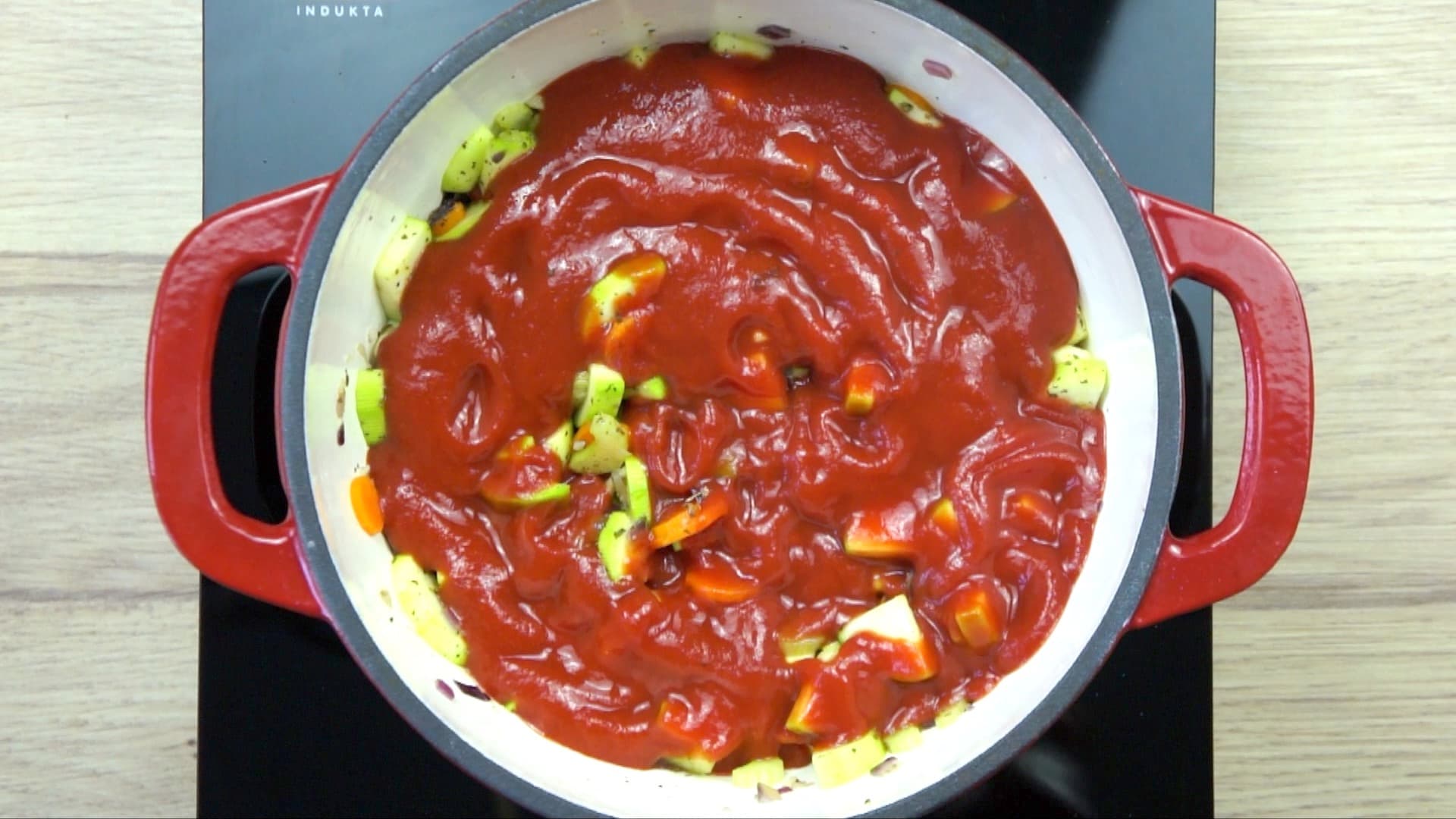 Chopped onion, garlic, zucchini, and sliced carrots covered in red tomato sauce in a white red enamelled Dutch oven.