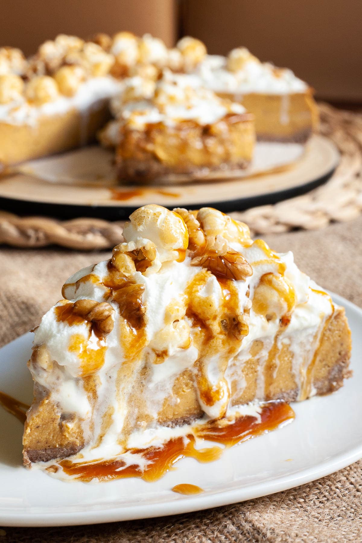 One slice of amber brown cheesecake with whipped cream, caramel sauce and chopped nuts on top is served on a white plate. The whole cake is in the background. 