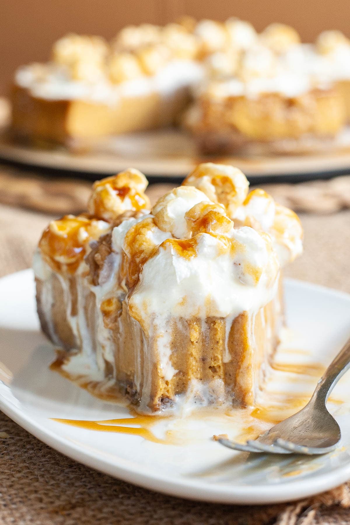 One slice of amber brown cheesecake with whipped cream, caramel sauce and chopped nuts on top is served on a white plate. One bite is missing from the front. The whole cake is in the background.