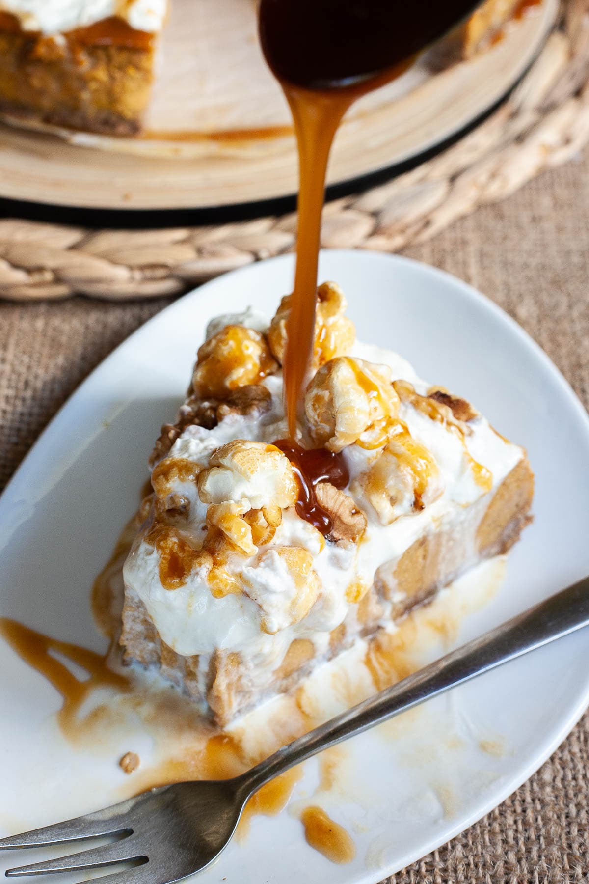 One slice of amber brown cheesecake with whipped cream, caramel sauce and chopped nuts on top is served on a white plate. More caramel sauce is flowing right on the top.