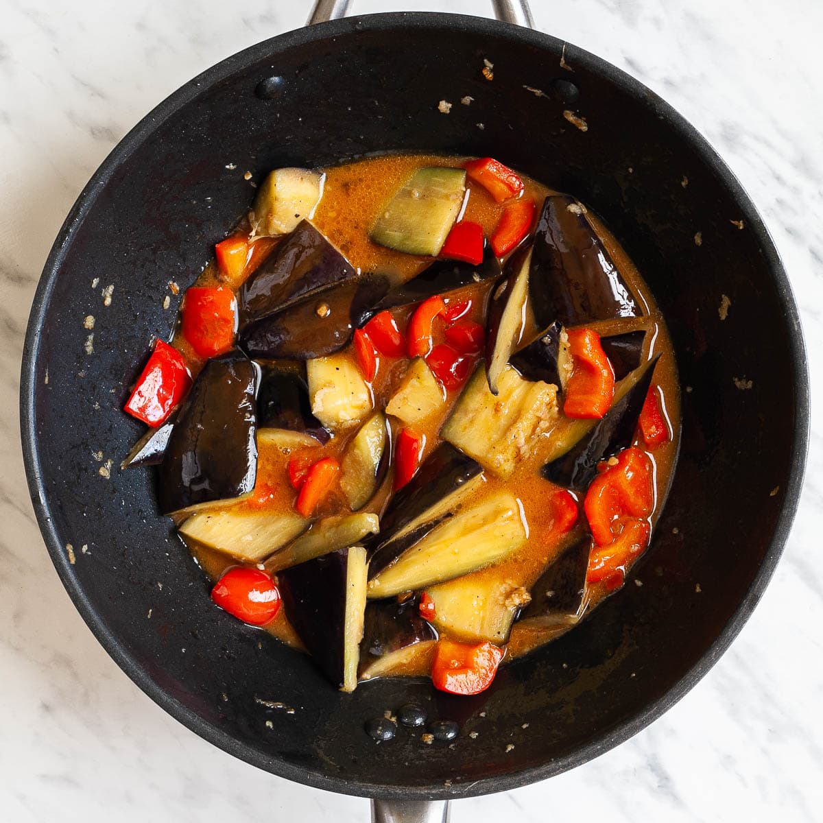 Eggplant slices and red pepper chunks in a wok are swimming in a brown sauce