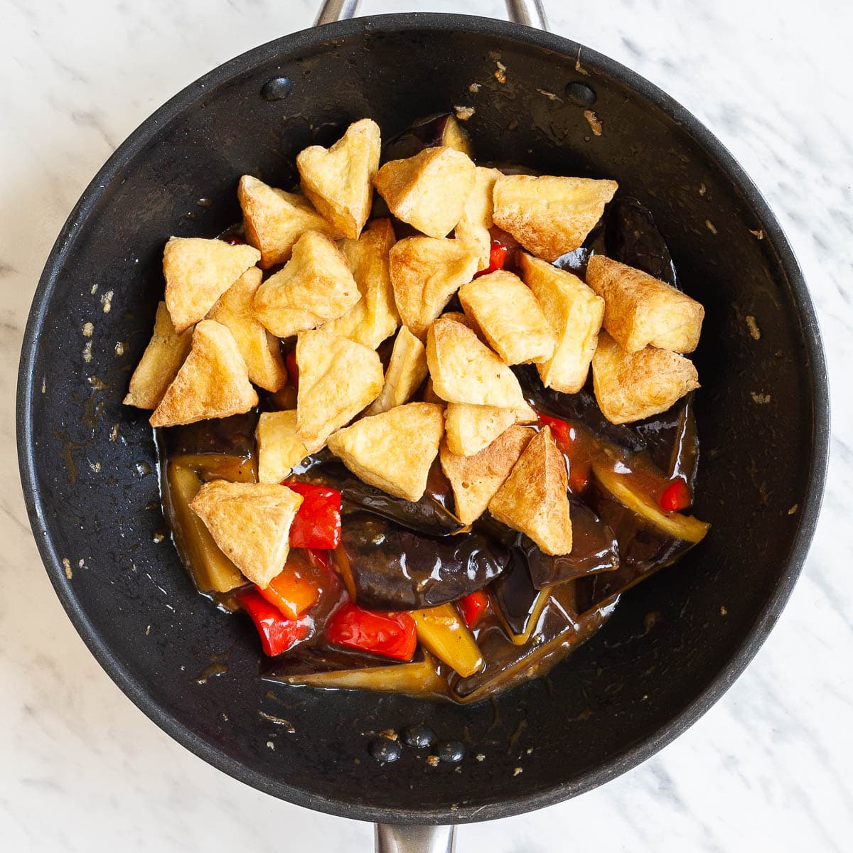 Puffy tofu triangles, eggplant slices and red pepper chunks in a wok are swimming in a brown sauce