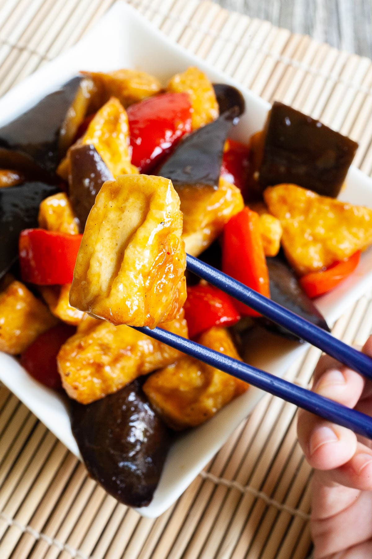 White rectangular plate full of large purple eggplant chunks, puffy brown glazed tofu and red peppers. Blue chopsticks are taking one tofu piece and holding it up in the air. 