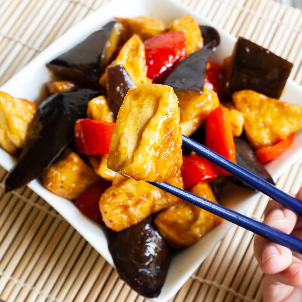 White rectangular plate full of large purple eggplant chunks, puffy brown glazed tofu and red peppers. Blue chopsticks are taking one tofu piece and holding it up in the air.