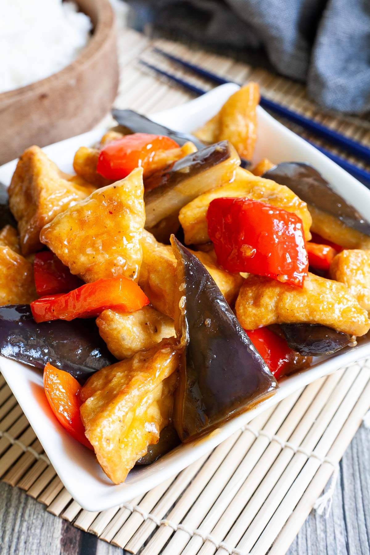 White rectangular plate full of large purple eggplant chunks, puffy brown glazed tofu and red peppers. Rice is served in a wooden bowl next to it.
