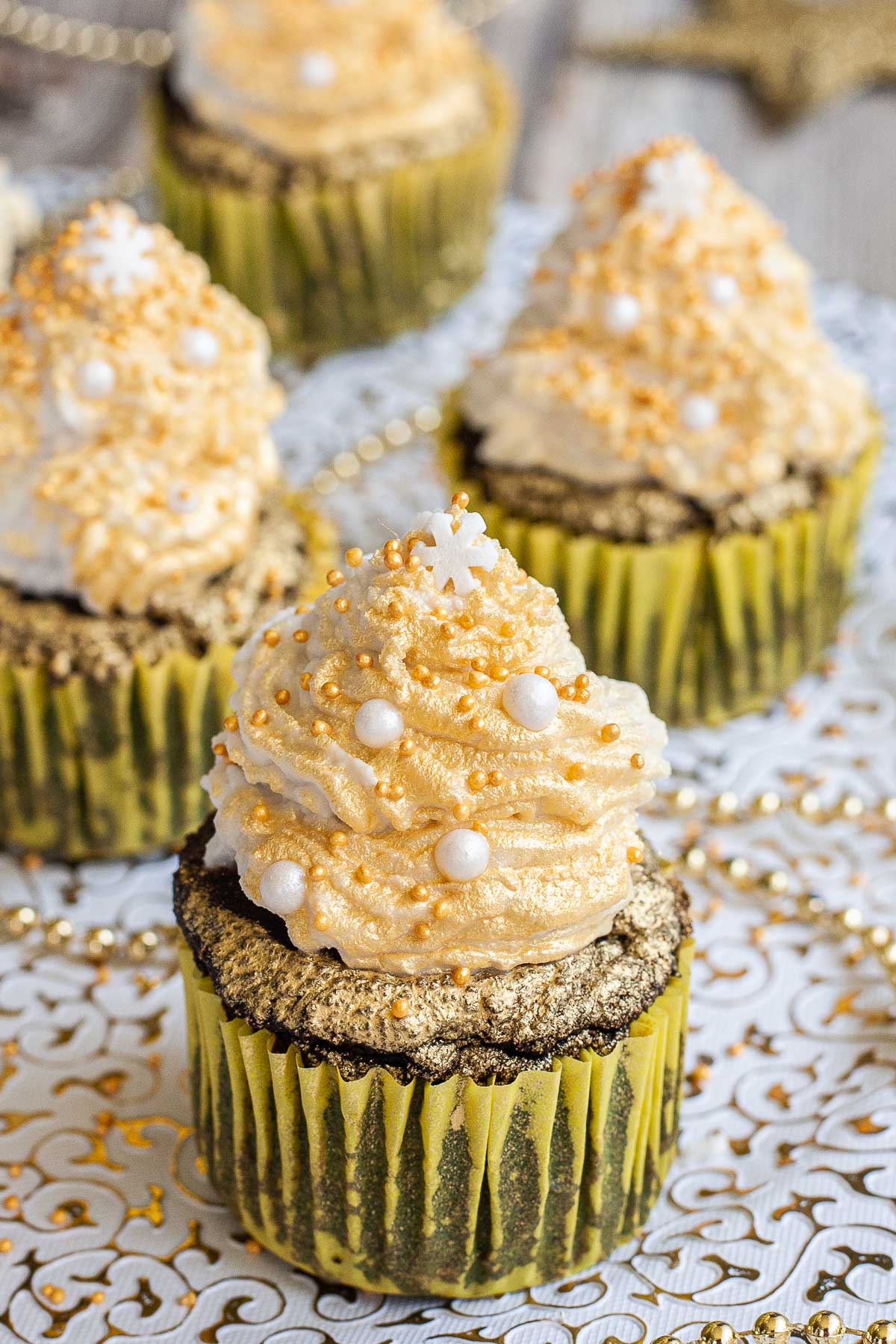4 dark brownie cupcakes in yellow cupcake liner topped with a white frosting sprayed with gold and sprinkled with gold and white pearl sprinkles and a snowflake one on top. 