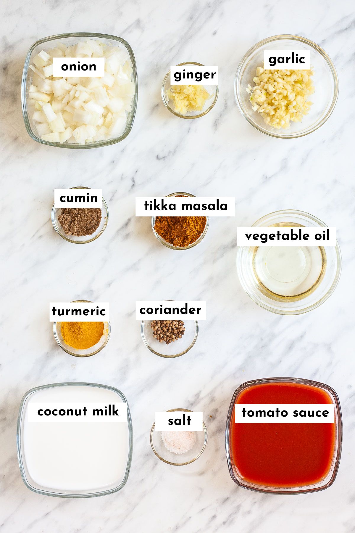 Ingredients of vegan tikka masala sauce placed in small glass bowls like chopped onion, minced garlic and ginger, different brown and yellow spices, red tomato sauce, thick white sauce, and oil