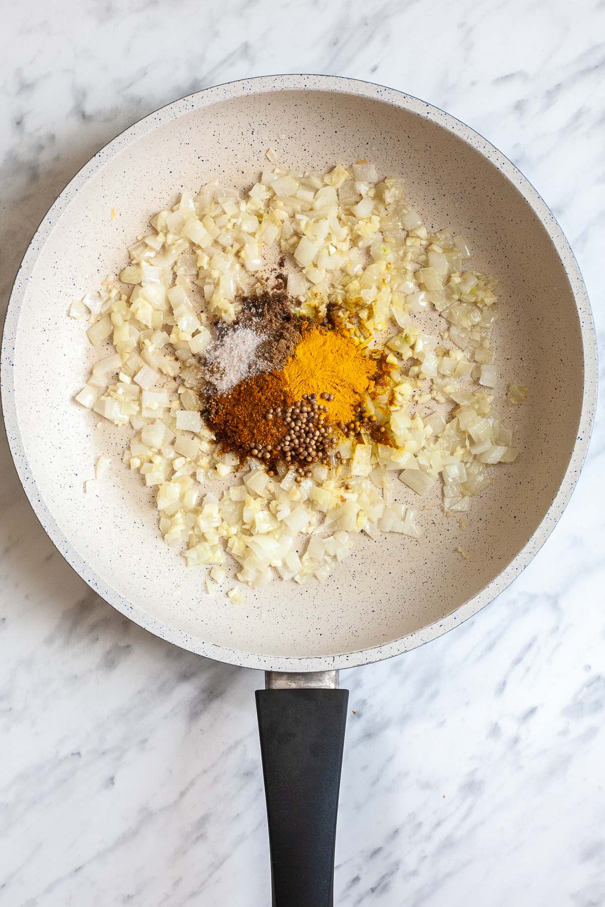 White frying pan with minced onion, garlic, and multiple heaps of spices. 
