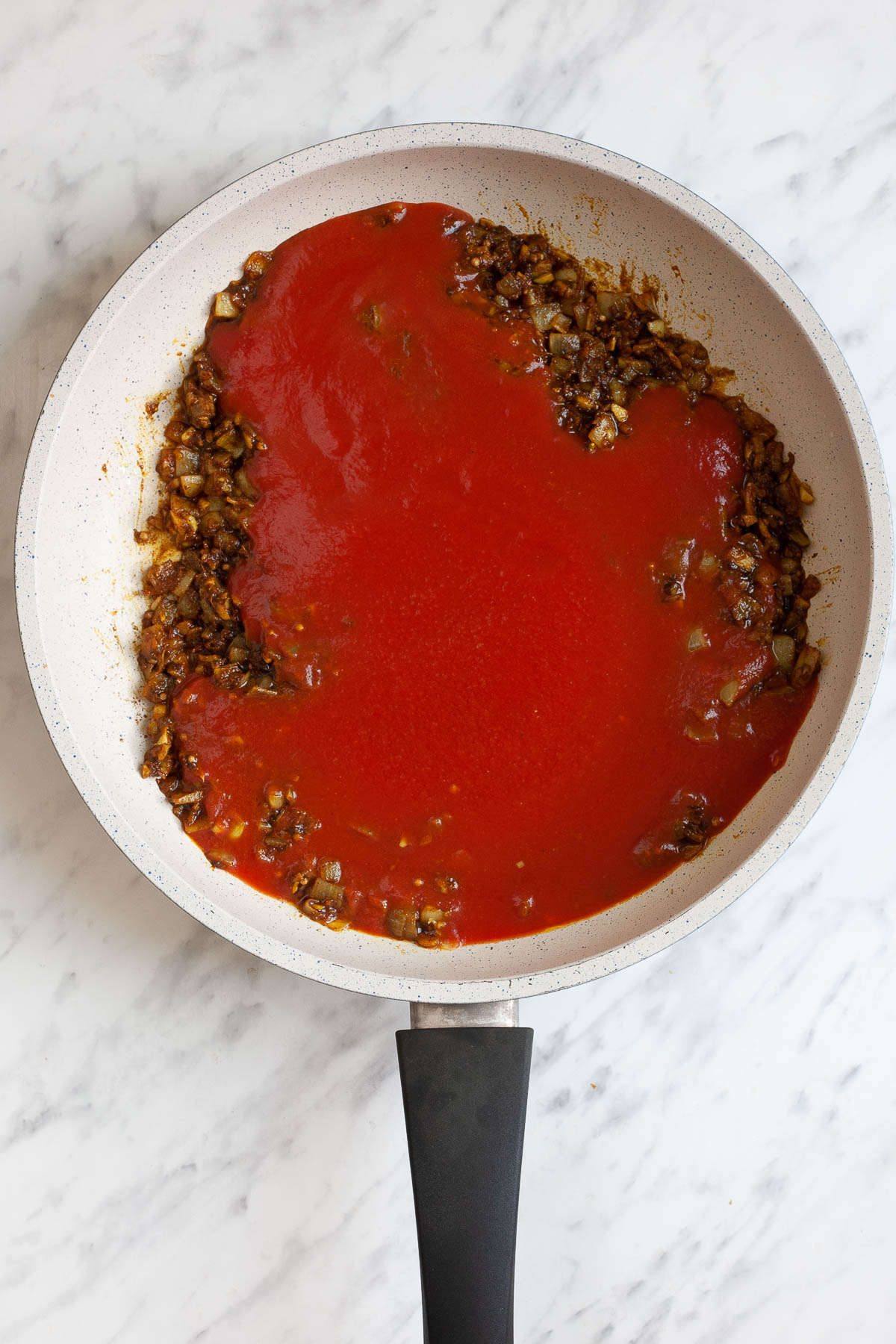 White frying pan with minced brown onion and red sauce.