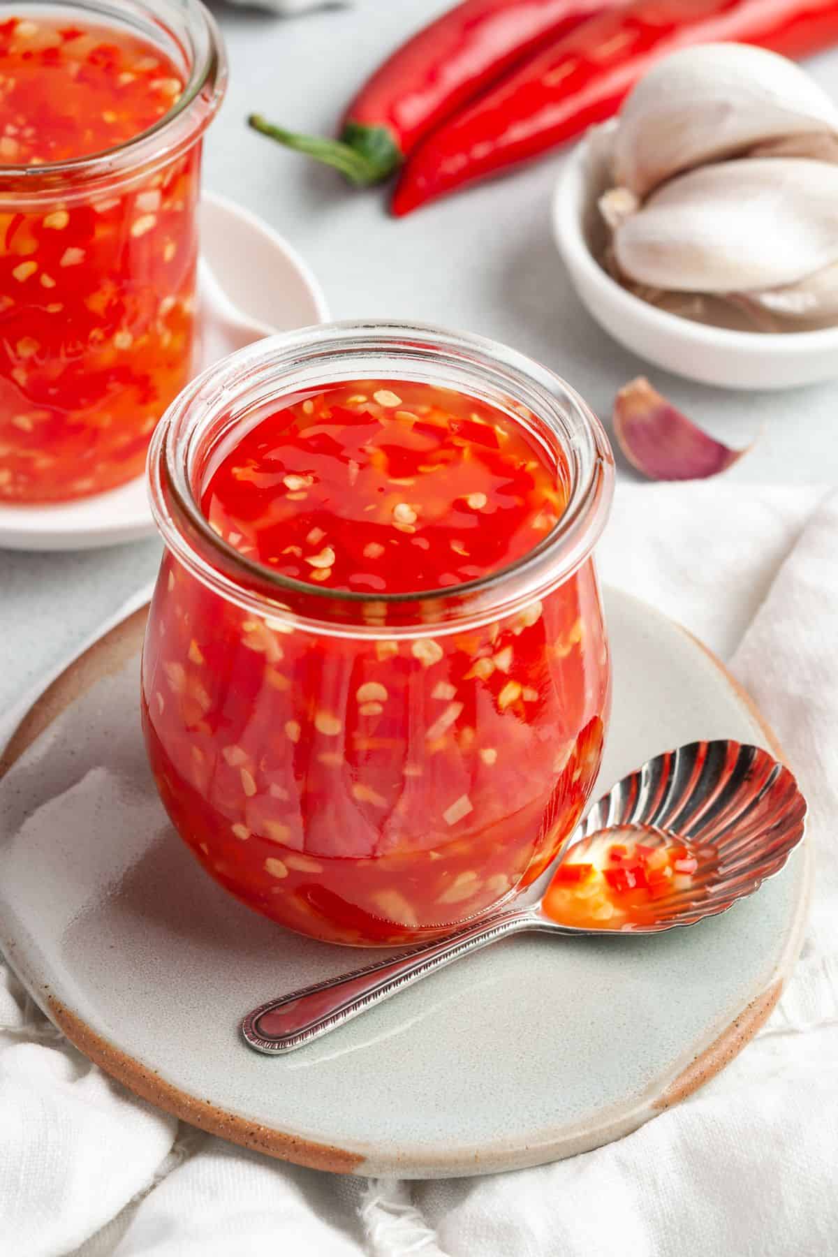 A chunky red sauce in a small glass jar placed on a light blue plate. A spoon with a bit of red sauce is place next to it.