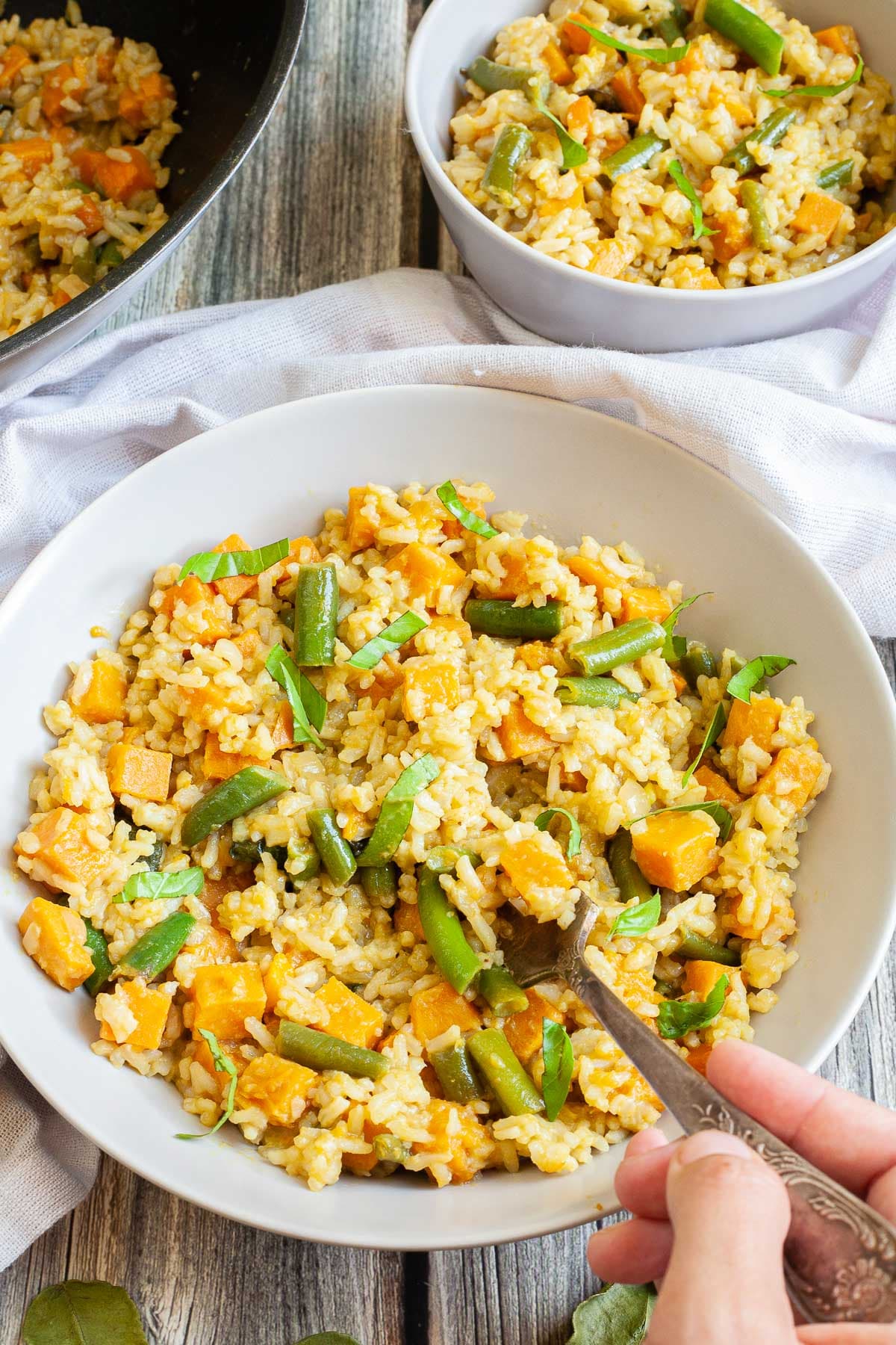 Two white bowls of fried rice in an orange sauce with sweet potato cubes and green bean pieces. A hand is holding a fork to take some.