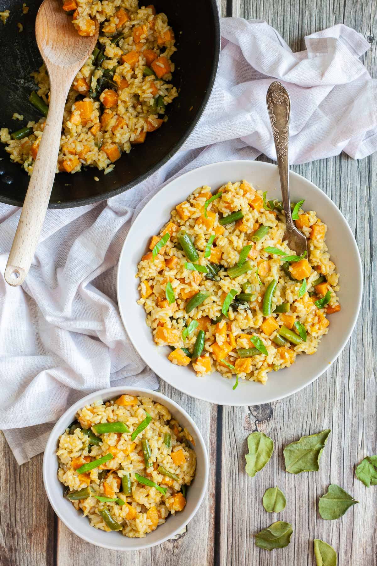 Two white bowls of fried rice in an orange sauce with sweet potato cubes and green bean pieces, A fork is placed inside the dish. Leftover green curry fried rice is in a wok next to the bowls.