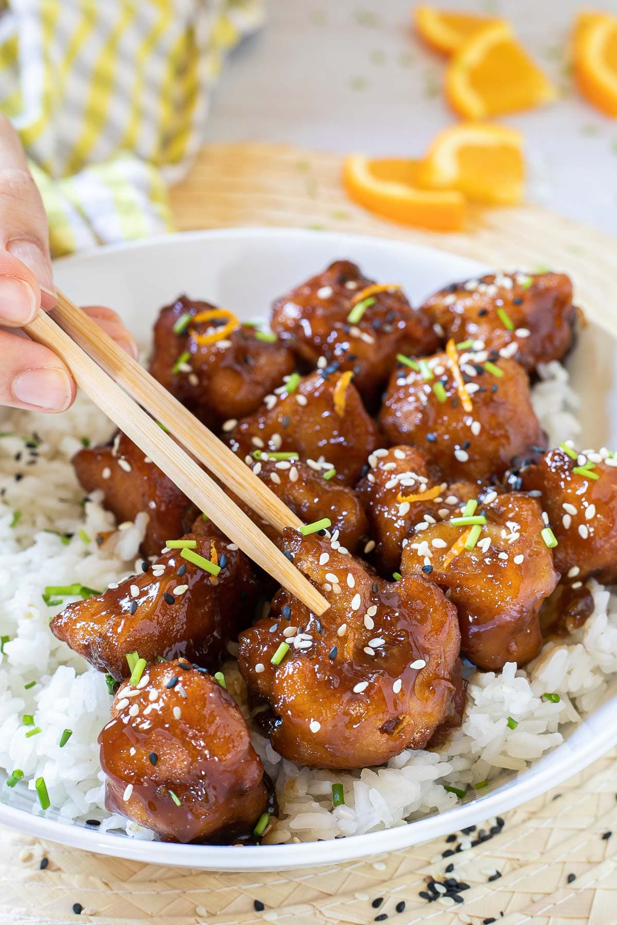 Sticky brown breaded cauliflower florets are served on top of rice in a white bowl. It is sprinkled with chopped chives and sesame seeds. A hand is holding chopsticks to take one.