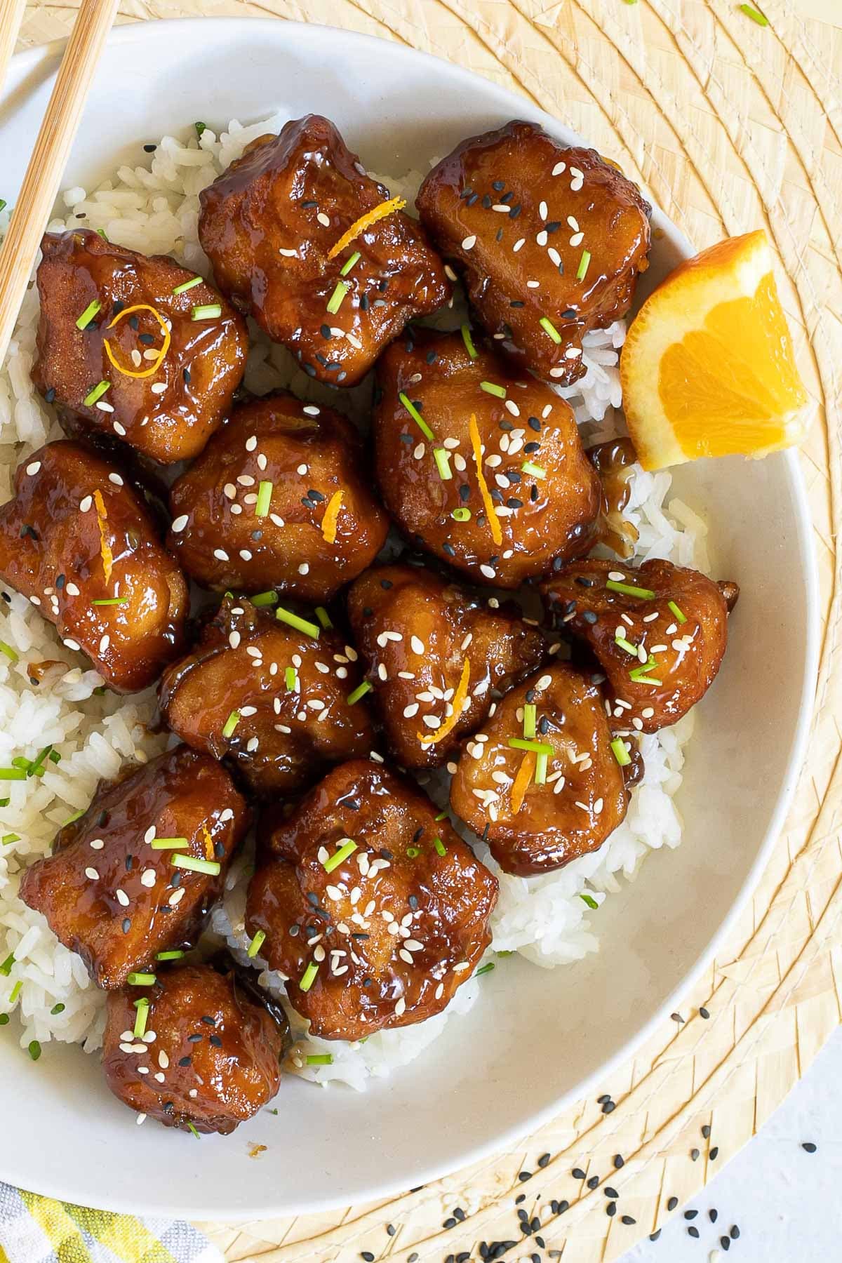 Sticky brown breaded cauliflower florets are served on top of rice in a white bowl. It is sprinkled with chopped chives and sesame seeds.