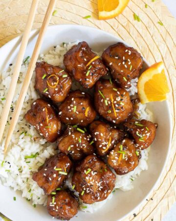 Sticky brown breaded cauliflower florets are served on top of rice in a white bowl. It is sprinkled with chopped chives and sesame seeds. Chopsticks are placed on top of the rice.