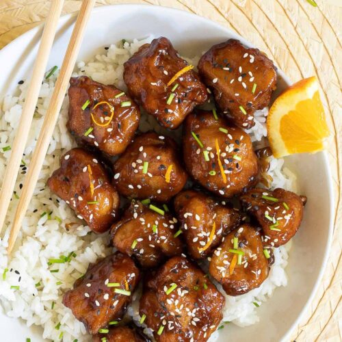 Sticky brown breaded cauliflower florets are served on top of rice in a white bowl. It is sprinkled with chopped chives and sesame seeds. Chopsticks are placed on top of the rice.
