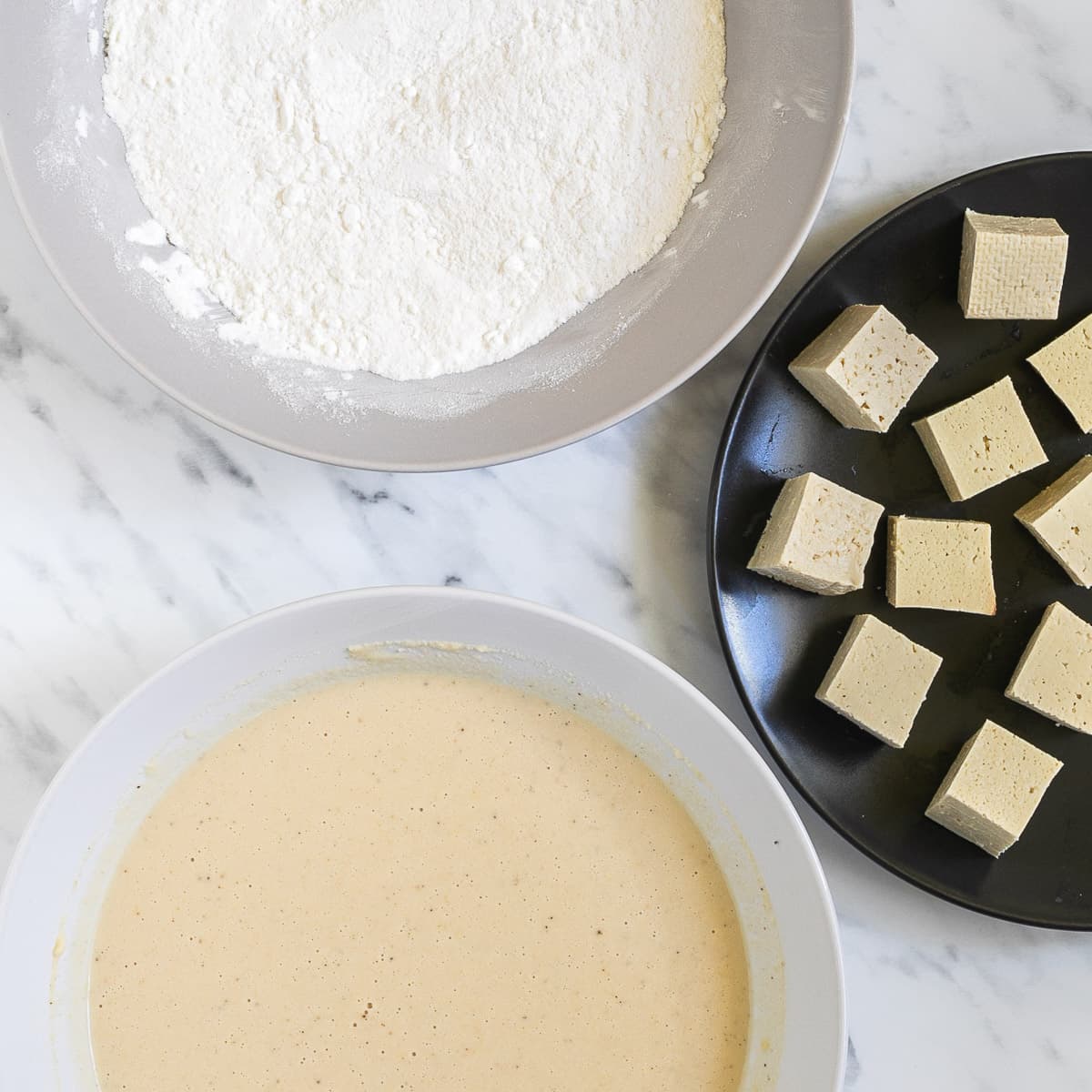 One white bowl of white flour, one white bowl of light yellow batter and one black bowl of tofu cubes.