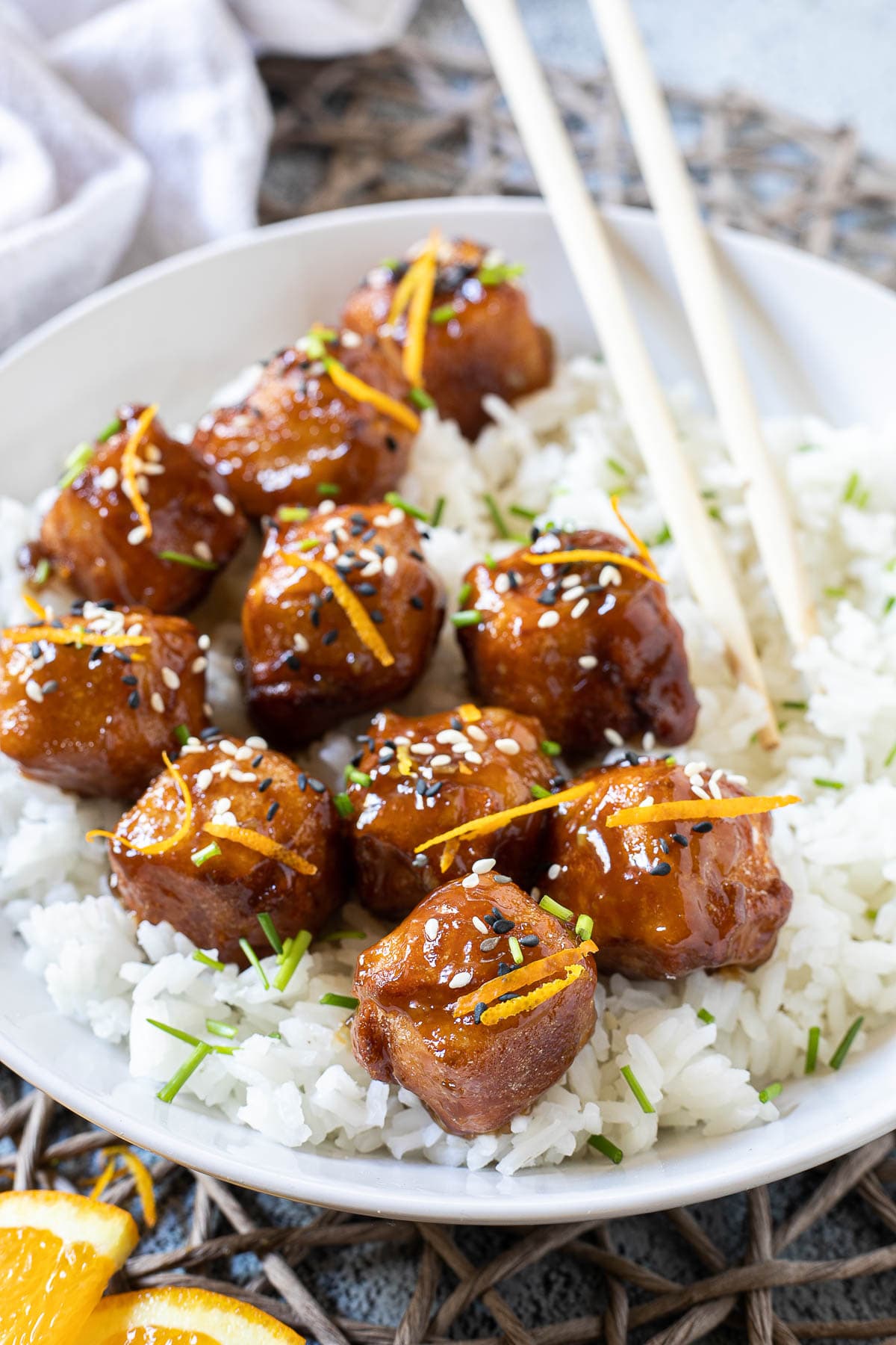 Sticky brown breaded tofu cubes are served on top of rice in a white bowl. It is sprinkled with chopped chives and sesame seeds. Chopsticks are placed on the rice.