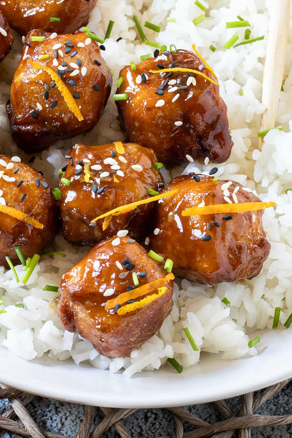 Sticky brown breaded tofu cubes are served on top of rice in a white bowl. It is sprinkled with chopped chives and sesame seeds.