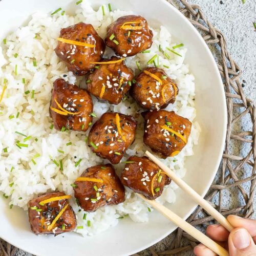 Sticky brown breaded tofu cubes are served on top of rice in a white bowl. It is sprinkled with chopped chives and sesame seeds. A hand is holding chopsticks to take one.