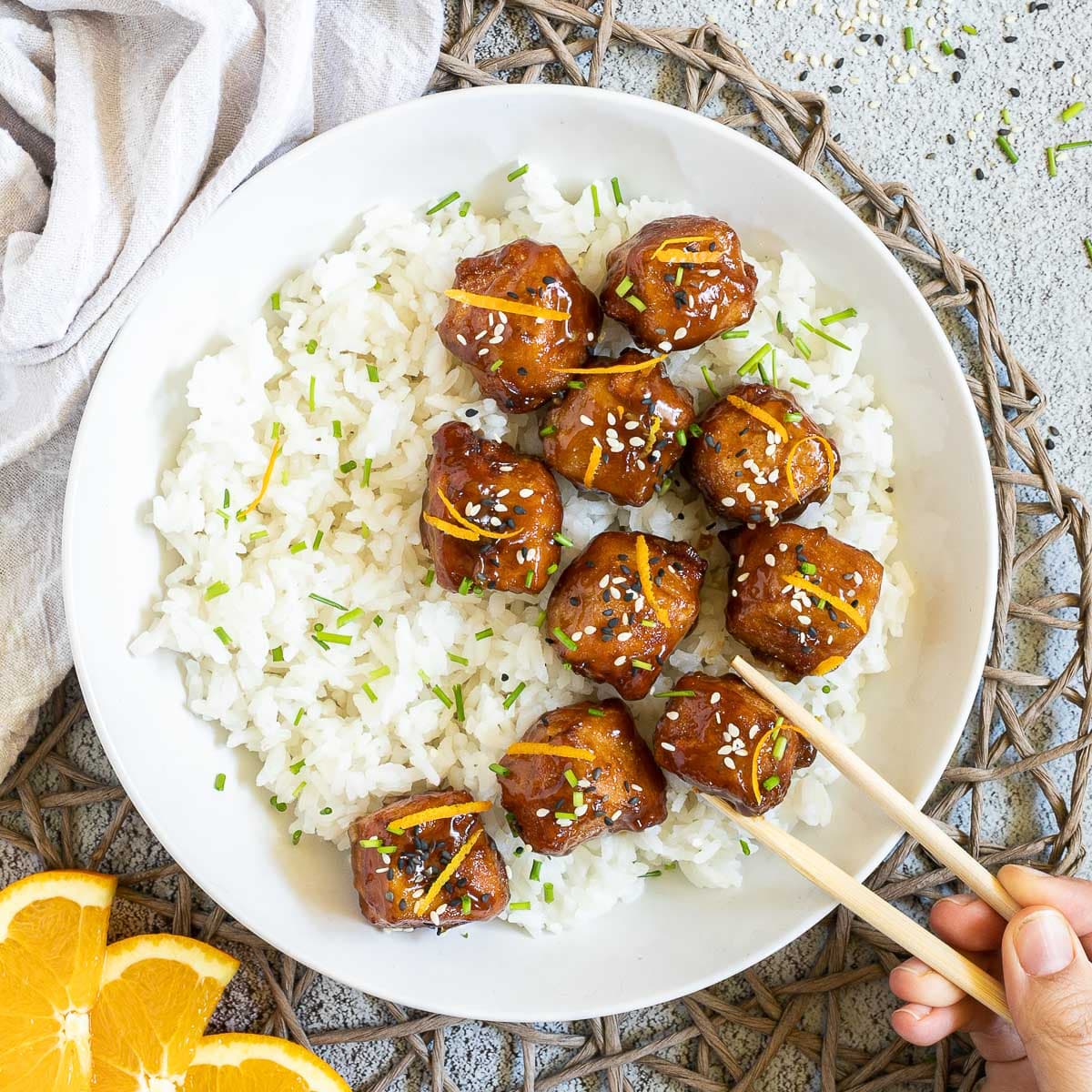 Sticky brown breaded tofu cubes are served on top of rice in a white bowl. It is sprinkled with chopped chives and sesame seeds. A hand is holding chopsticks to take one.