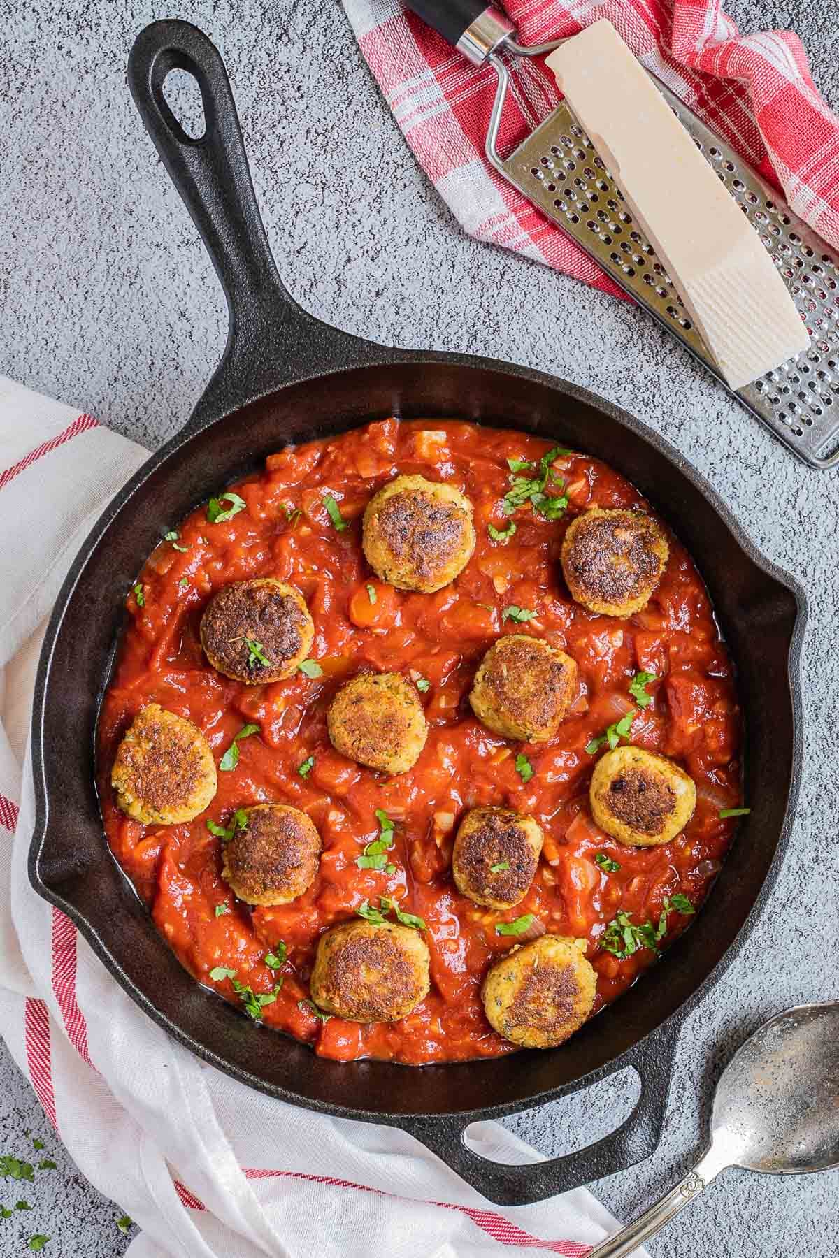 Black cast iron skillet with red marinara sauce and crispy brown tofu meatballs. It is sprinkled with chopped fresh green herbs. Vegan Parmesan cheese is next to it on top of a cheese grater. 