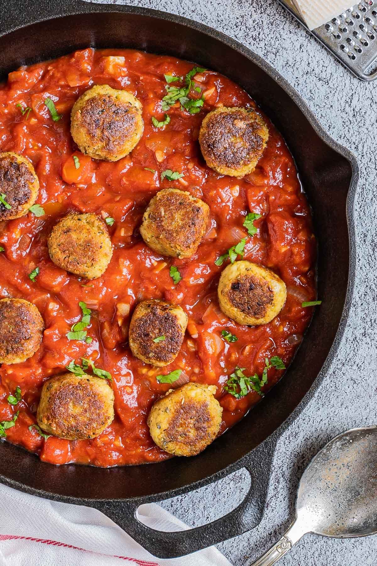 Black cast iron skillet with red marinara sauce and crispy brown tofu meatballs. It is sprinkled with chopped fresh green herbs.