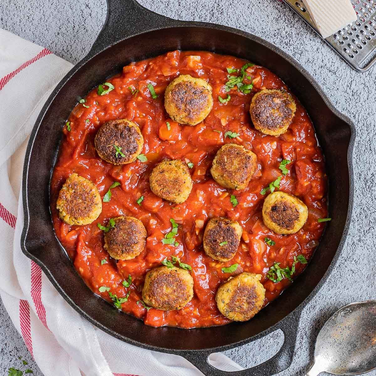 Black cast iron skillet with red marinara sauce and crispy brown tofu meatballs. It is sprinkled with chopped fresh green herbs. Vegan Parmesan cheese is next to it on top of a cheese grater.