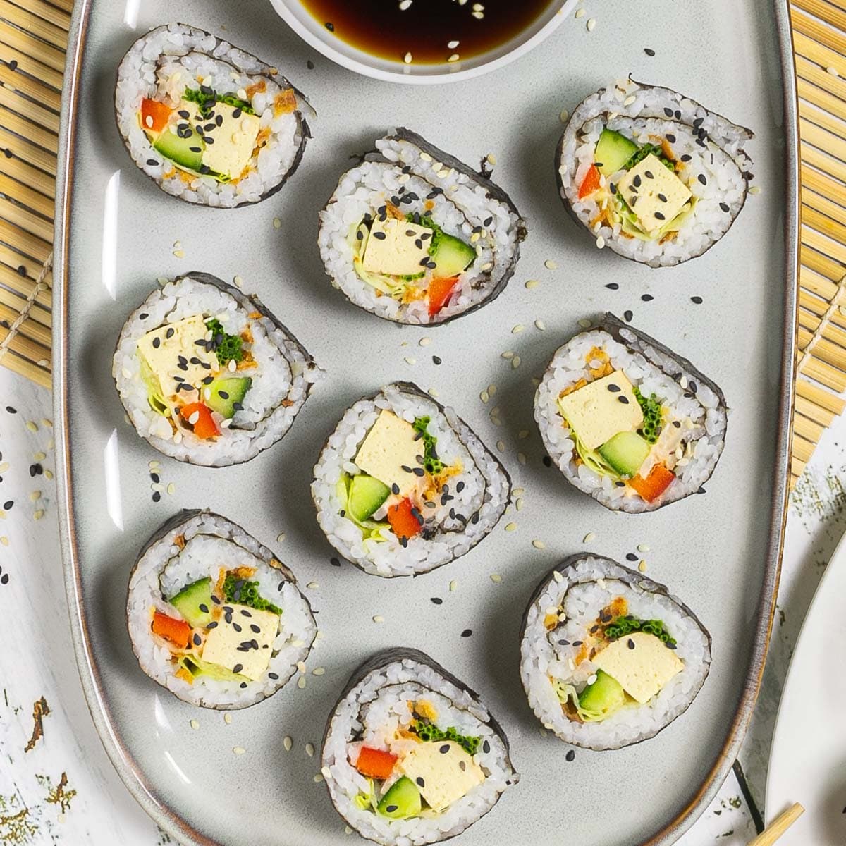 An oval, light blue plate with 9 tofu sushi rolls with colorful filling wrapped in dark seaweed sheets. Served with a small bowl of soy sauce and sprinkled with dark and white sesame seeds.