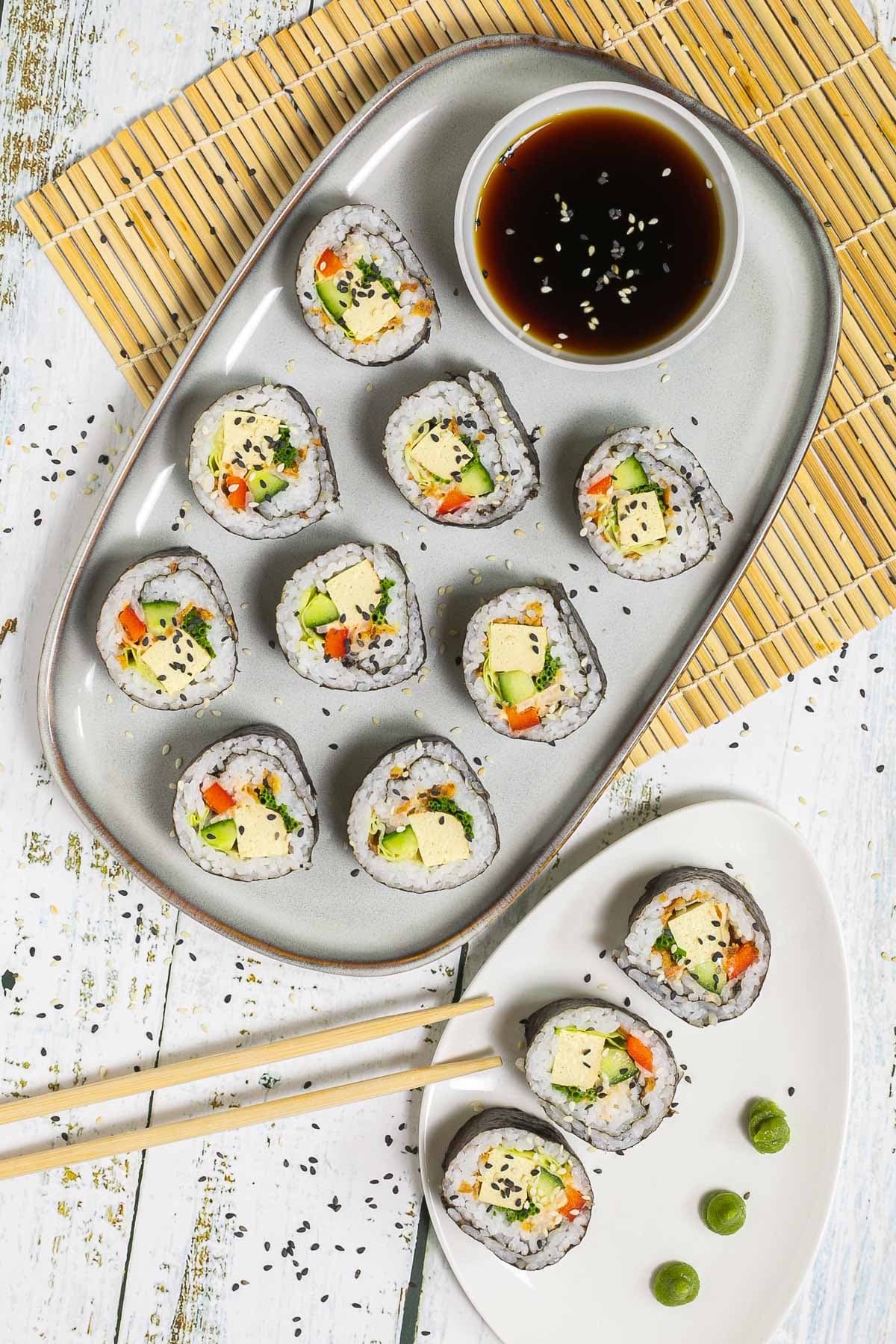 An oval, light blue plate with 9 tofu sushi rolls with colorful filling wrapped in dark seaweed sheets. Served with a small bowl of soy sauce and sprinkled with dark and white sesame seeds.
